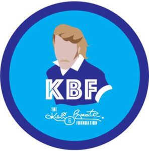 Circular logo with a faceless illustration of a person with light hair and mustache, wearing a blue shirt. Text reads "KBF" and "The King Beatle Foundation" with a stylized signature.